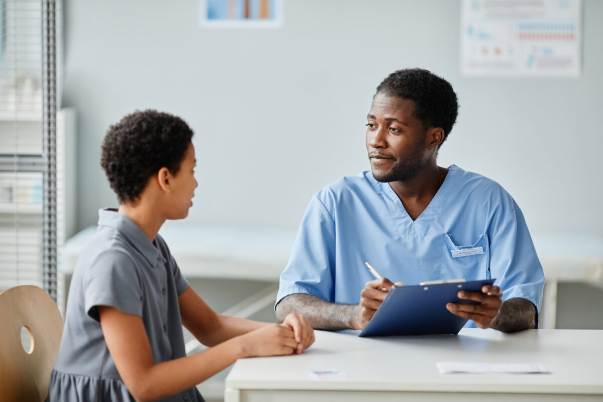Portrait of young African-American doctor talking to teenage girl in medical clinic, copy space