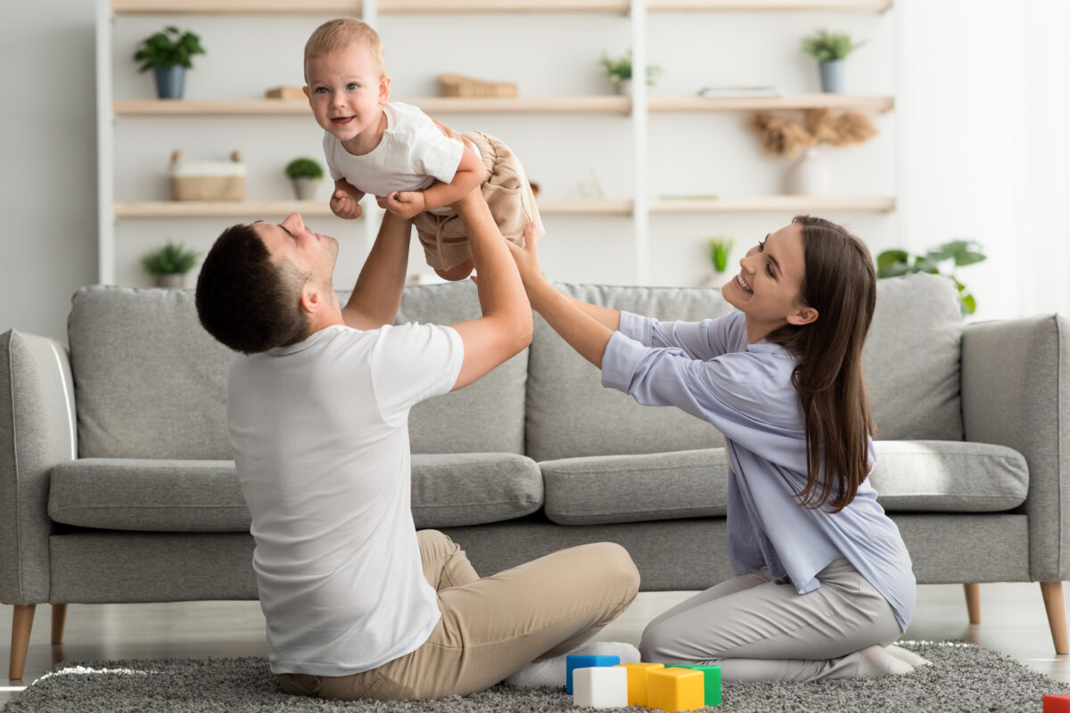 Young Parents Having Fun With Their Adorable Toddler Son At Home, Loving Mom And Dad Playing With Cute Infant Child And Laughing, Enjoying Spending Time Together, Dad Lifting Excited Baby, Free Space
