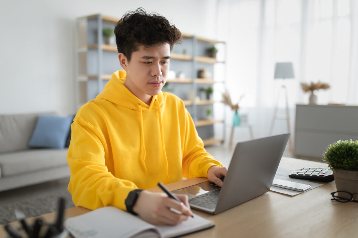 Portrait of serious focused young Asian man in yellow hoodie sitting at desk, using pc typing on keyboard and writing research taking notes, working online at home office